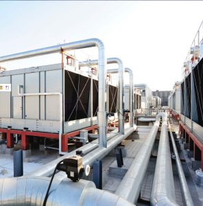 A different angle of the industrial rooftop cooling tower plant system showing multiple large metal cooling units connected by an extensive network of silver pipes running along the concrete surface, with buildings visible in the background.
