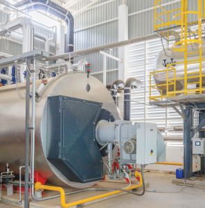 A large industrial boiler installed in a modern plant room, surrounded by metal piping, insulation, and a yellow service platform under bright natural light.