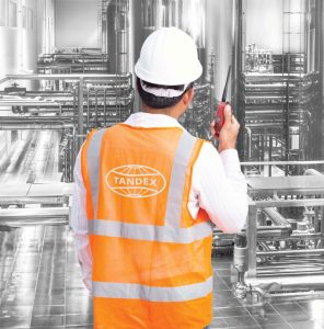 A Tandex technician wearing a white hard hat and orange safety vest with the Tandex logo uses a two-way radio while inspecting stainless steel plant equipment and pipework inside an industrial facility.