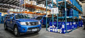Blue Tandex service vehicles parked inside the company warehouse in front of racking filled with blue chemical drums and industrial storage containers.