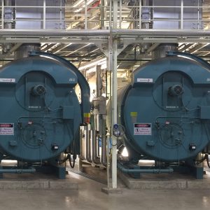 Two large industrial boilers viewed from a slightly different angle inside a customer facility, showing the same twin boiler setup with surrounding pipes, gauges, and warning signage.