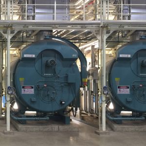 Two large industrial boilers installed side by side on a customer site, surrounded by metal piping and safety signage within a clean, well-lit plant room.