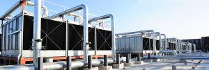 A large industrial cooling tower plant system installed on a rooftop, featuring multiple rectangular cooling tower units connected by extensive silver metal piping under a clear blue sky.
