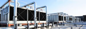 A large industrial cooling tower plant system installed on a rooftop, featuring multiple rectangular cooling tower units connected by extensive silver metal piping under a clear blue sky.
