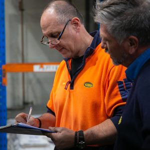 A Tandex factory hand in an orange uniform and a technician in a navy jumper reviewing an audit checklist together on a clipboard inside the warehouse.