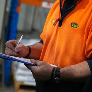 Close-up view of a Tandex factory worker wearing an orange uniform with the company logo, holding a clipboard and pen while completing a checklist inside the warehouse.