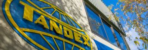 Close-up of the Tandex logo on the front of the company’s office building, with reflections in the windows and tree branches visible above.