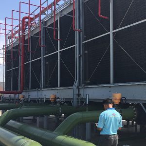 A Tandex representative wearing a light blue company shirt stands in front of a large industrial cooling tower system, inspecting equipment and piping. The system features tall black tower units, red overhead pipes, and green water lines beneath them.