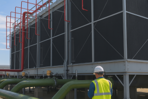 A Tandex service technician wearing a hard hat and high-visibility vest stands in front of a large industrial cooling tower system. The cooling towers feature red and green piping and metal framework, set outdoors under a bright sky.