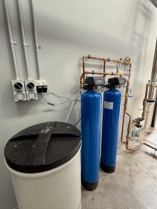 Industrial duplex softener unit featuring two blue resin tanks connected by copper pipework, with a white brine tank on the left used for system regeneration.