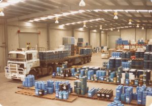 Historic photo from 1991 showing the Tandex factory being set up, with a truck unloading blue chemical drums and containers inside the warehouse.