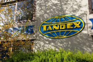 The Tandex logo displayed on the front wall of the company building, partially framed by green plants and tree branches.