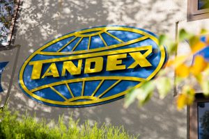 Closer view of the Tandex logo mounted on the front wall of the company building, surrounded by greenery and tree shadows.