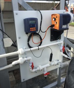 Wall-mounted wastewater monitoring panel with inline sensor, control display, and automatic chemical dosing pump connected to pipework and isolation valves for water quality control.