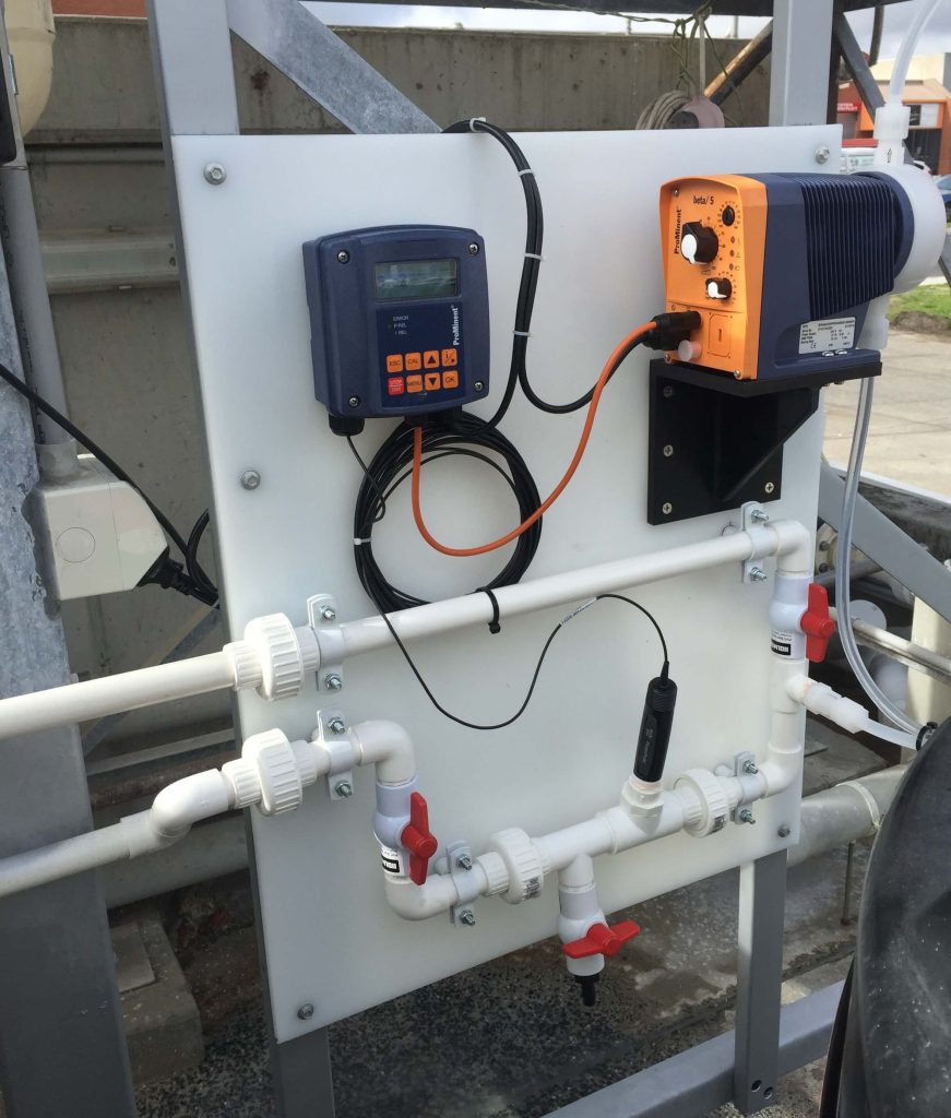 Wastewater monitoring and automatic dosing system Wall-mounted wastewater monitoring panel with inline sensor, control display, and automatic chemical dosing pump connected to pipework and isolation valves for water quality control.