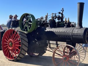Close-up of a black steam traction engine with red wheels and a green flywheel in motion, operated by two people during a heritage machinery event under clear blue skies.