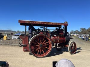 Red steam traction engine labeled “Horton & Morris Threshing Contractors” driving along a dirt track at a heritage machinery event, with an operator standing at the controls and spectators watching in the background.