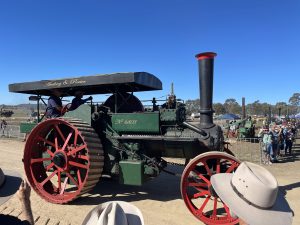 Green and red steam traction engine labeled “Aveling & Porter No. 6803” driving along a dirt track at a heritage machinery event, with operators on board and spectators watching from the side.