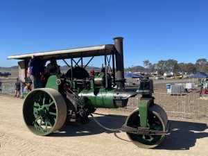 Green steam roller with canopy driven along a dirt track during a heritage machinery event, with operators standing on the platform and spectators watching under clear blue skies.