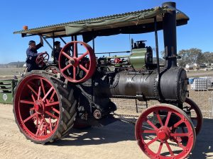 Vintage steam traction engine with a green boiler and red wheels driving along a dirt track during a heritage machinery event, with two operators under a canopy and spectators in the background.