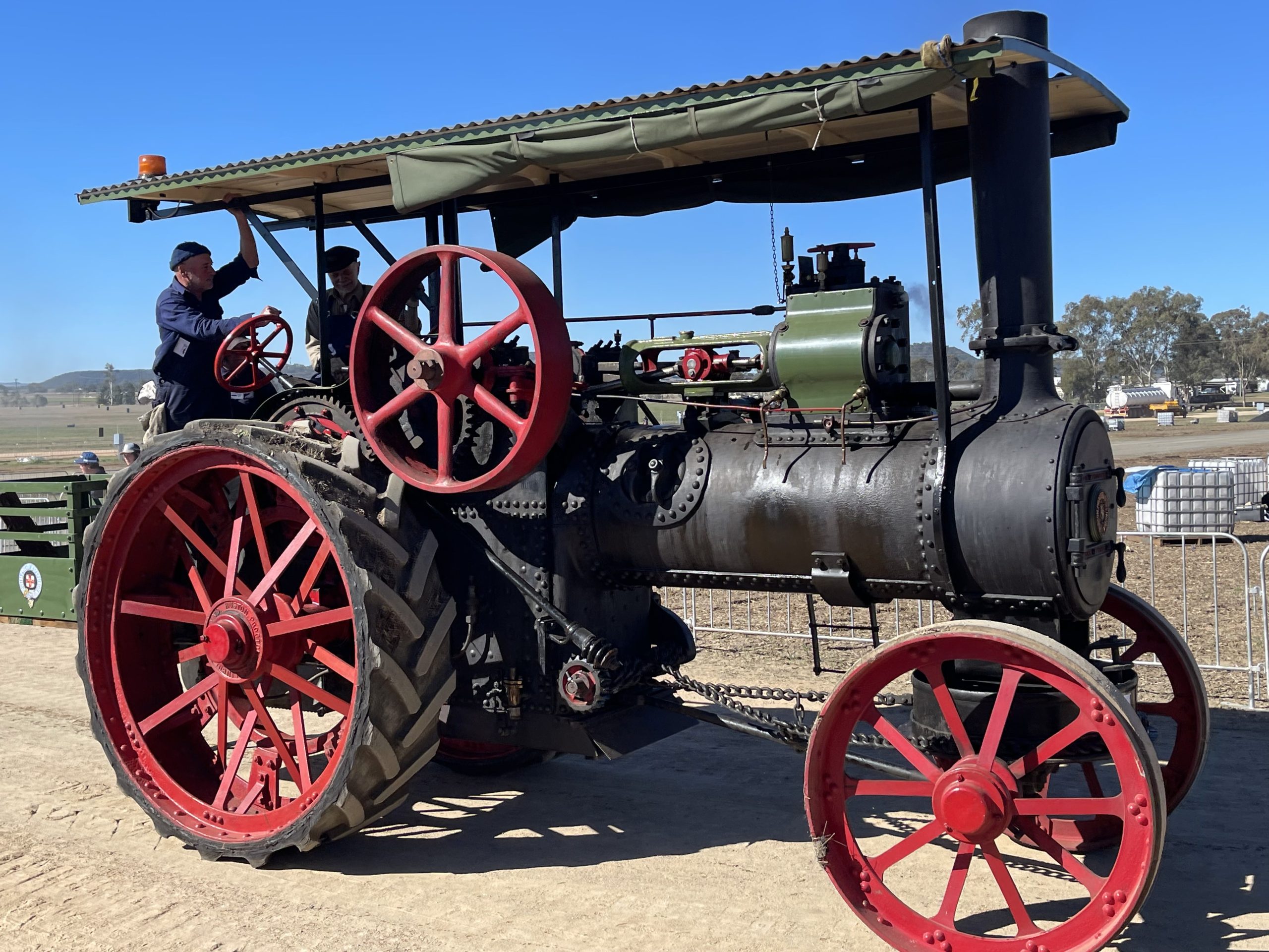 Vintage steam traction engine with a green boiler and red wheels driving along a dirt track during a heritage machinery event, with two operators under a canopy and spectators in the background.