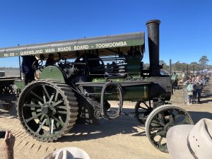 Dark green steam traction engine labeled “Queensland Main Roads Board – Toowoomba No.128” driving along a dirt track with operators on board and spectators watching at a heritage machinery event.