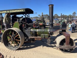 Olive green steam roller labeled “Carr & Company” driving along a dirt track at a heritage machinery event, with operators on board and spectators watching nearby.