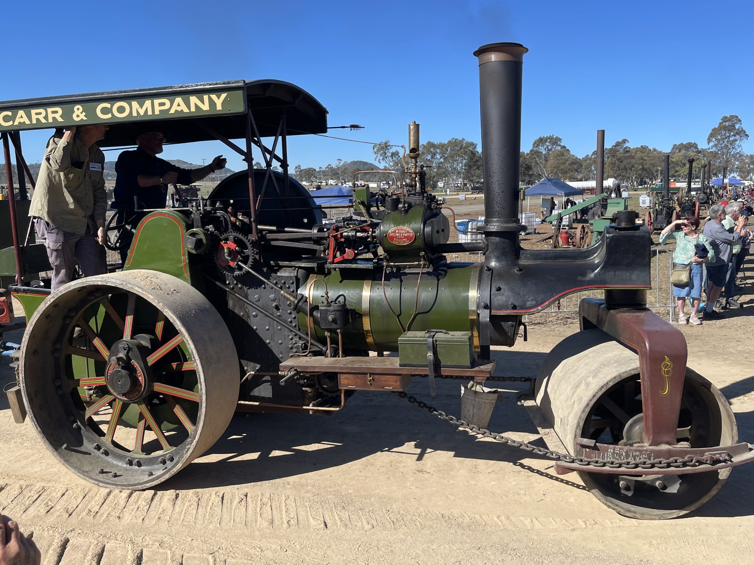 Olive green steam roller labeled “Carr & Company” driving along a dirt track at a heritage machinery event, with operators on board and spectators watching nearby.