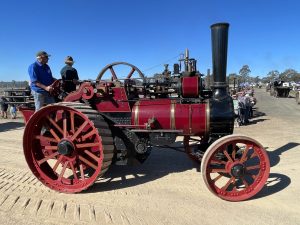 Red and black steam traction engine with gold trim driving along a dirt track at a heritage machinery rally, with operators standing on the platform and spectators watching in the background.