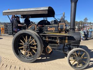 Vintage black and gold steam traction engine named “Sooty” driving along a dirt track with operators on board and spectators watching at a heritage machinery event.