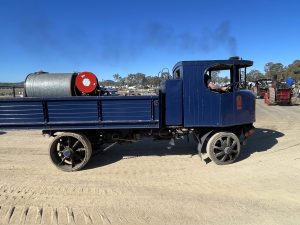 Blue steam-powered lorry with an open cab and metal drum load driving along a dirt track at a heritage machinery rally, with other steam vehicles and spectators in the background.