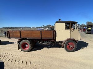 Beige and brown vintage steam lorry with a timber tray driving along a dirt track during a heritage machinery event, with spectators and other steam vehicles in the background.