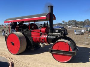 Bright red and black vintage steam roller driving along a dirt track during a heritage machinery event, with spectators and tents visible in the background under clear blue skies.