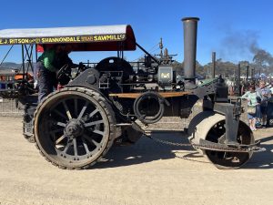 Black steam roller with a red canopy reading “E.J. Ritson Shannondale Steam Sawmill Co” driven along a dirt track during a heritage machinery rally.