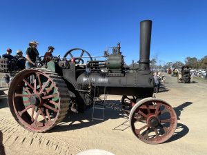 Steam traction engine driving along a dirt track during a heritage machinery rally, with spectators watching under a clear blue sky.