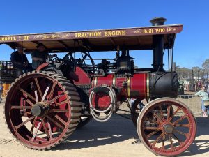 A beautifully restored red and black Burrell 7 horsepower traction engine built in 1910, with large spoked wheels and a canopy labelled “Three Speed Traction Engine.” The steam engine is displayed at an outdoor heritage rally with onlookers in the background under clear blue skies.