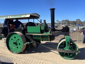 A vintage green steam road roller with a canopy reading “Ruston & Hornsby Ltd.” The machine features large metal rollers and a tall black chimney, displayed outdoors at a heritage machinery rally under clear blue skies.
