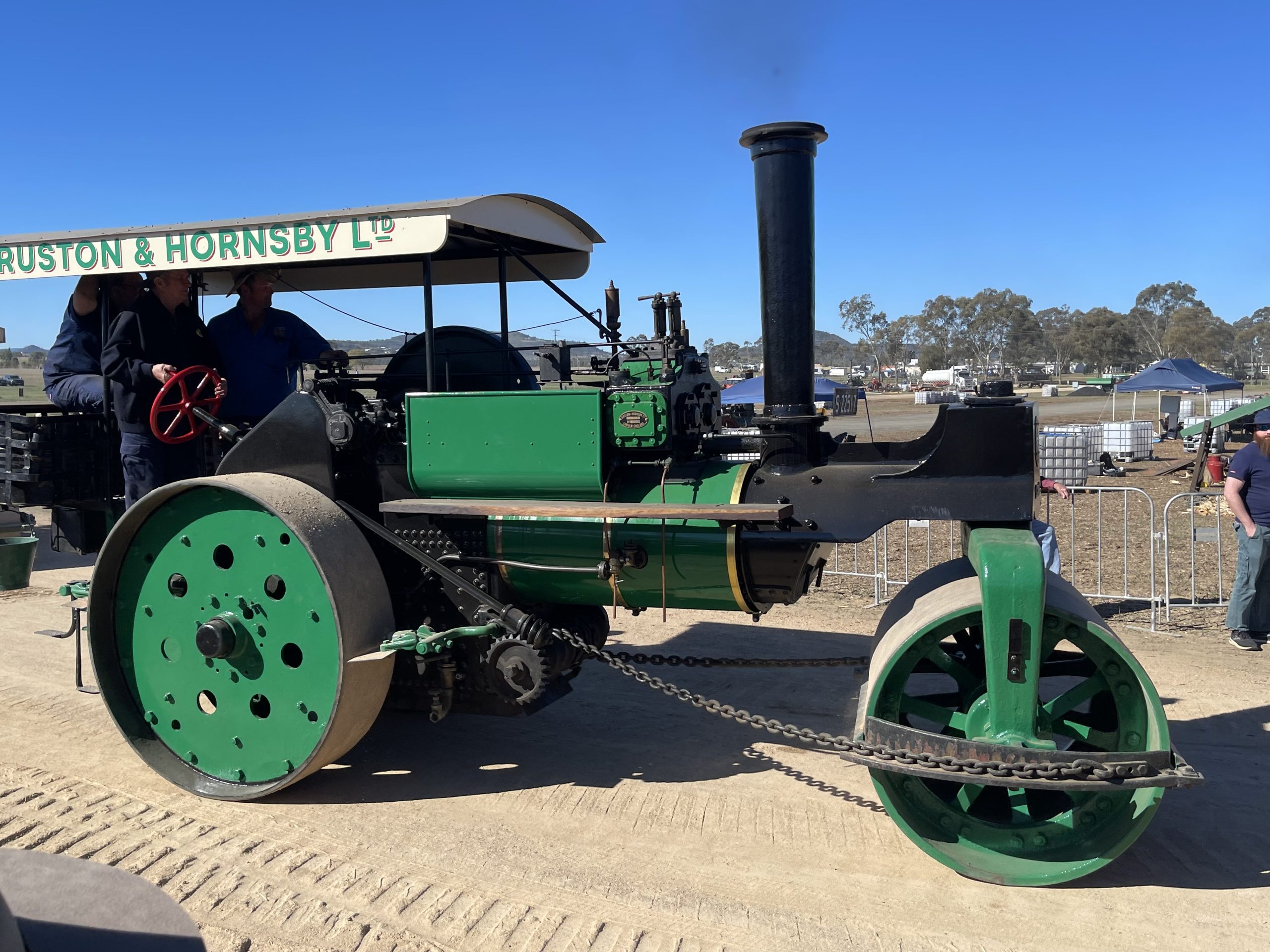A vintage green steam road roller with a canopy reading “Ruston & Hornsby Ltd.” The machine features large metal rollers and a tall black chimney, displayed outdoors at a heritage machinery rally under clear blue skies.