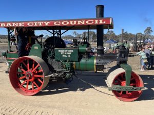 A restored green and black steam road roller with red wheels and a canopy reading “Brisbane City Council.” The roller is labelled “Dept. of Works No. 16” and is displayed outdoors at a heritage machinery rally with people watching in the background.