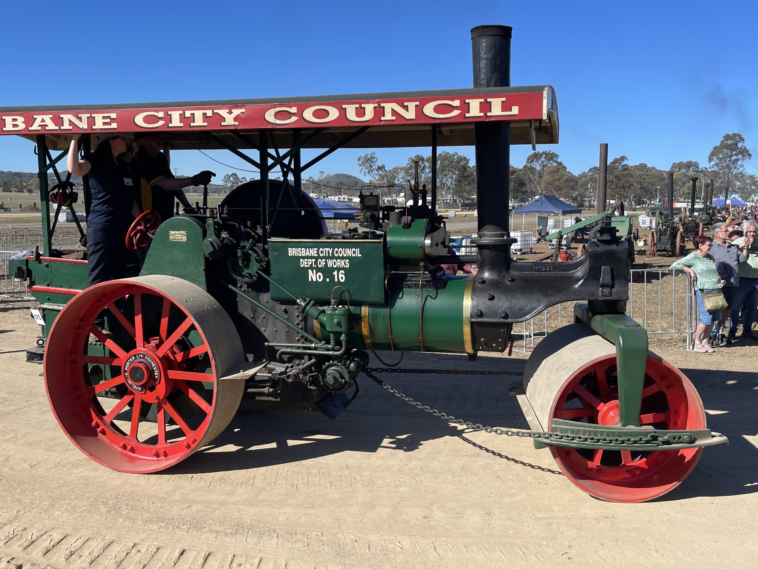 A restored green and black steam road roller with red wheels and a canopy reading “Brisbane City Council.” The roller is labelled “Dept. of Works No. 16” and is displayed outdoors at a heritage machinery rally with people watching in the background.
