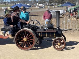 A man and child ride a miniature steam traction engine with large red wheels and brass fittings at an outdoor rally. Other participants stand nearby under blue tents and the area is fenced with metal barriers.