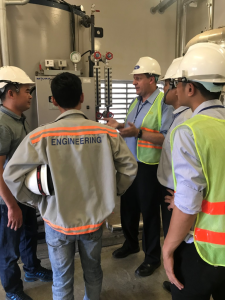 A Tandex operations manager discusses maintenance procedures with a group of international agents during a training visit, all wearing safety helmets and high-visibility vests in an industrial boiler room.