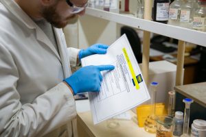 A Tandex laboratory technician wearing safety glasses and gloves examines a procedural document while performing a chemical assessment in the lab.