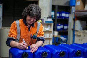 A Tandex factory worker in an orange high-visibility uniform marking blue chemical drums with a yellow marker as part of the manufacturing process.