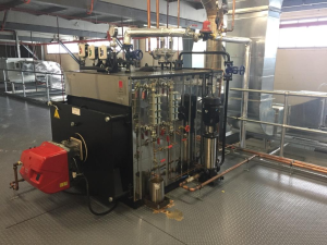 An industrial boiler system installed on a customer site, featuring a red burner unit, multiple gauges, copper piping, and stainless-steel components within a metal-floored plant room.