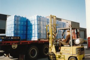 A Tandex factory worker operating a forklift to load stacks of blue chemical drums onto a truck outside the warehouse in 1991.