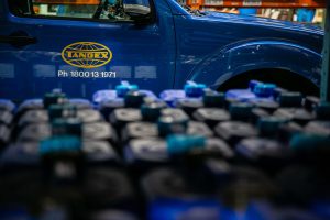 Blurred view of blue Tandex chemical drums on storage racking in the foreground, with a Tandex service vehicle and company logo visible on the door in the background.