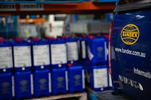 Close-up of a blue Tandex service vehicle with the company logo and website address visible on the tailgate, parked inside the factory. Rows of blue chemical drums are stacked on pallets in the blurred background, showing the warehouse environment.