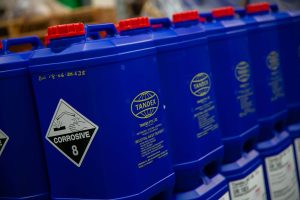 Row of blue Tandex chemical drums with yellow company logos and corrosive safety labels, stored in an industrial facility.