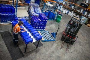 Overhead view of a Tandex factory worker in a high-visibility uniform labelling blue chemical drums on a workbench, surrounded by pallets, drums, and a forklift in the manufacturing area.